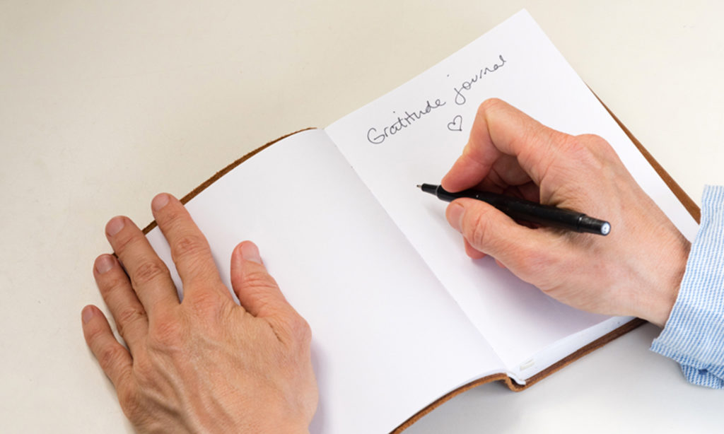 Hands hold a pen and write in a gratitude journal sitting on a white table